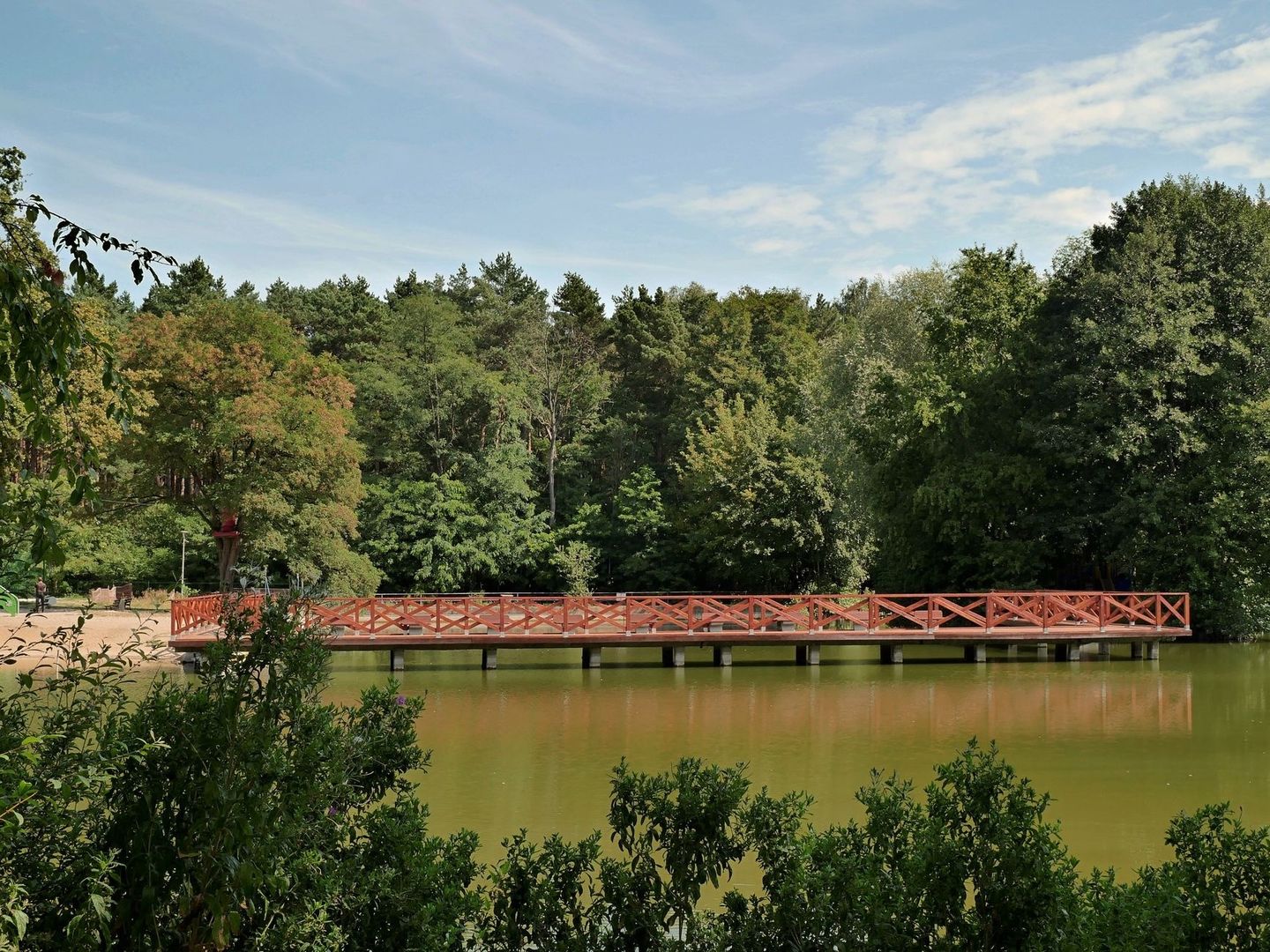 Röd träbro över en stilla sjö i en grönskande parkmiljö